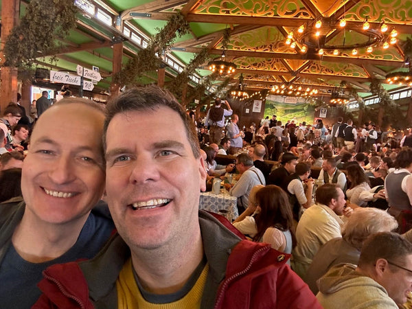 Two men posing for a selfie in a crowded indoor setting with decorative ceiling lights.